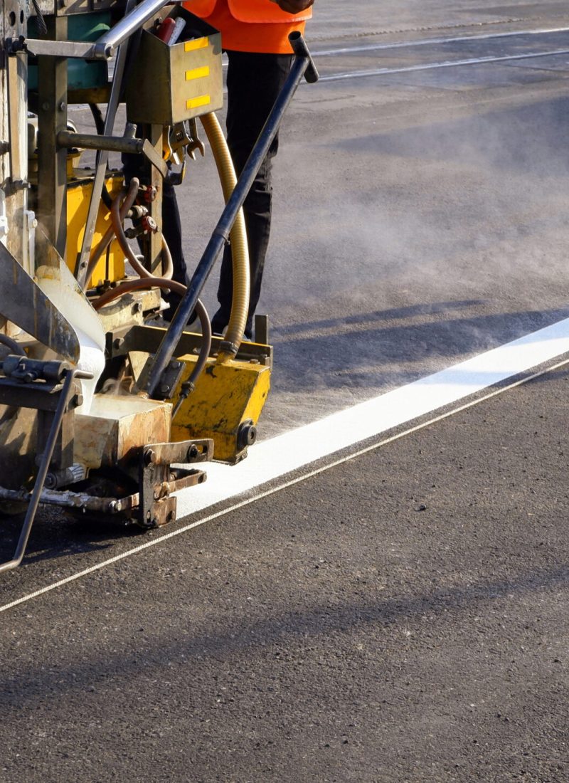 Low section of road worker with thermoplastic spray road marking machine working to paint traffic white  line with steam and railway track crossing on asphalt road surface